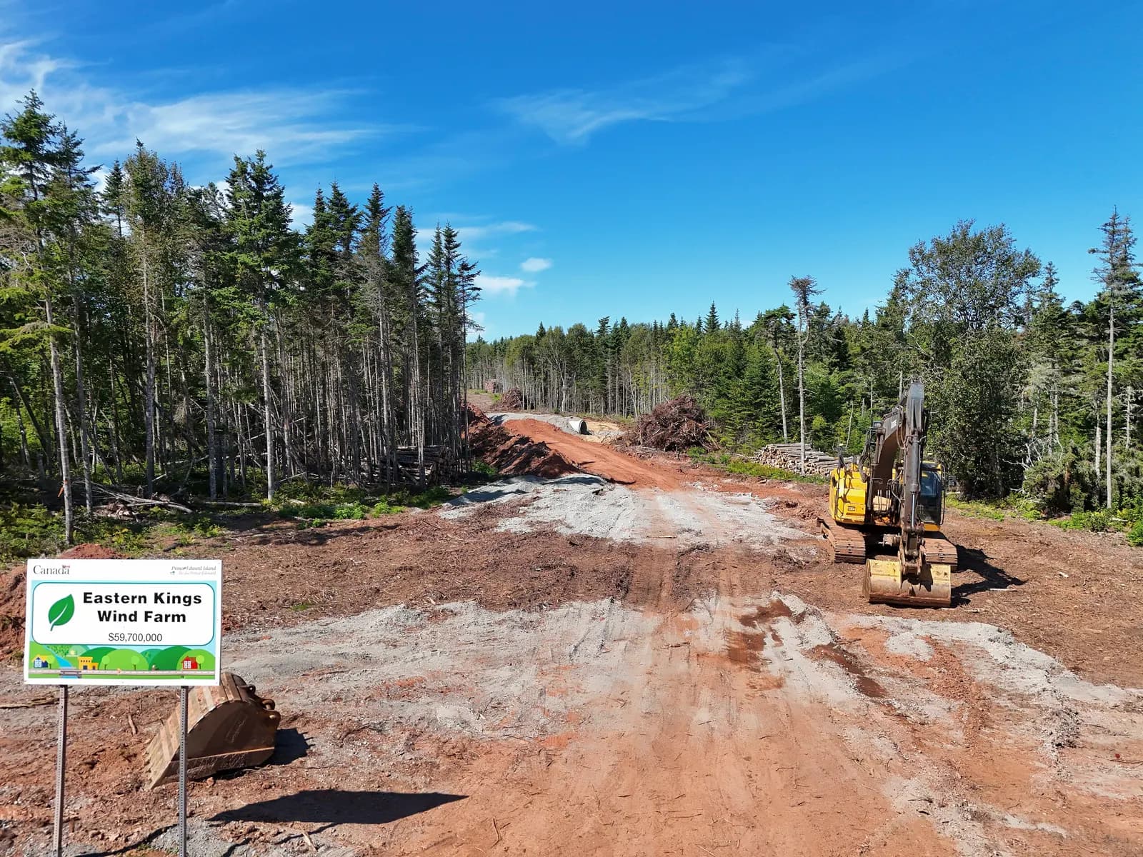 Cleared red-soil access road through spruce forest with an excavator, log piles and an Eastern Kings Wind Farm project sign