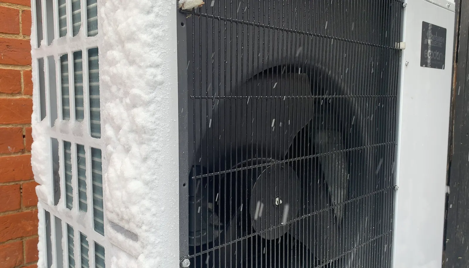 Outdoor air-source heat pump unit with snow and frost on its coil, beside a brick wall